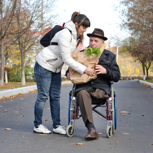 Senior man confined to a wheelchair by a leg amputation being helped with groceries by a woman carer in the street as they return from doing his shopping